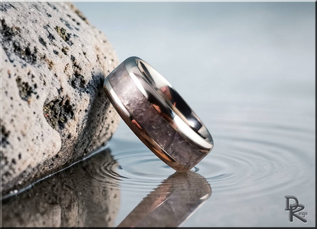 DR Rings titanium ring with inlay resting on rock by calm water with ripple reflections