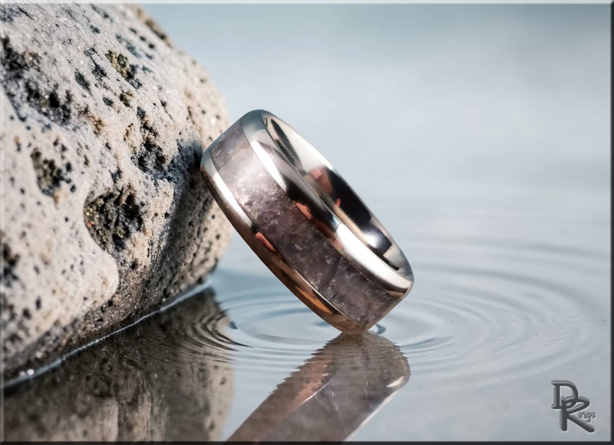 DR Rings titanium ring with inlay resting on rock by calm water with ripple reflections