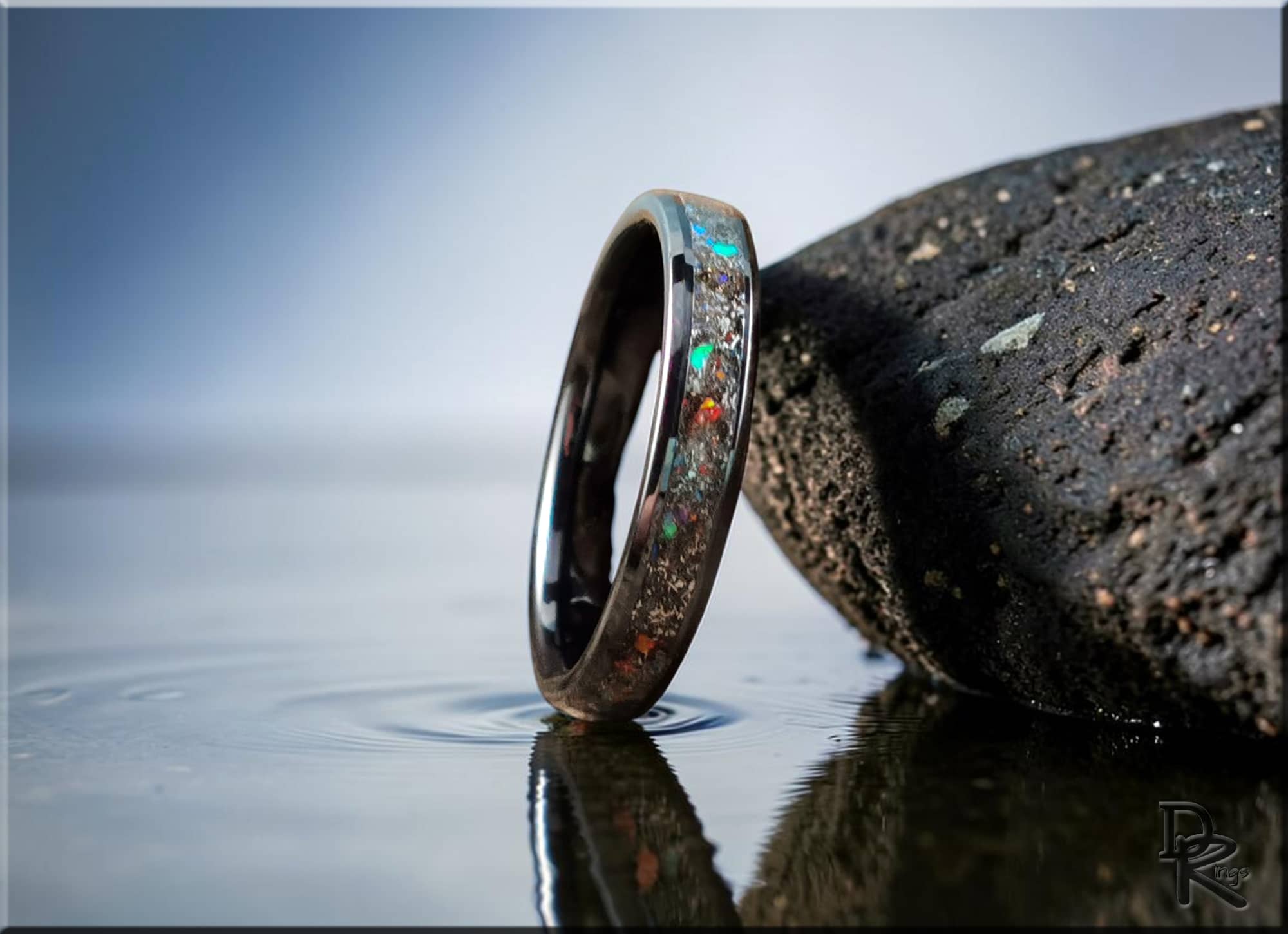 Ceramic ring with inlay featuring colorful opal accents displayed near a textured black rock