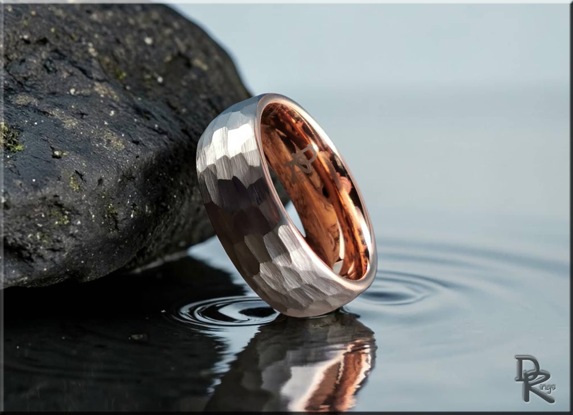 Hammered tungsten ring with rose gold core reflecting on water next to a textured rock by DR Rings