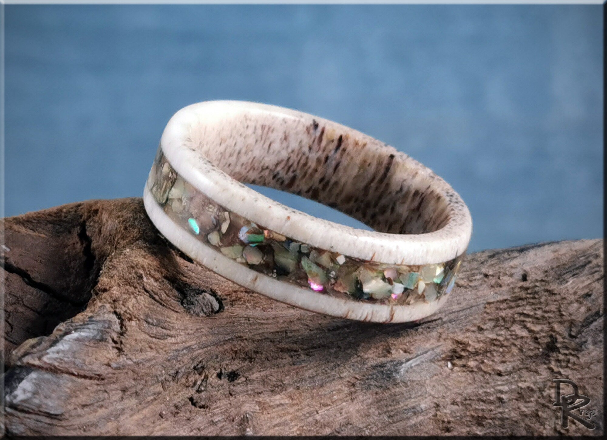 DR Rings antler ring with abalone inlay resting on rustic wood with blue background