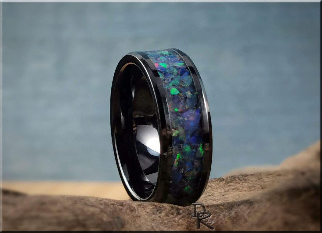 Black ceramic ring with opal inlay featuring vibrant blue and green hues on natural wood background
