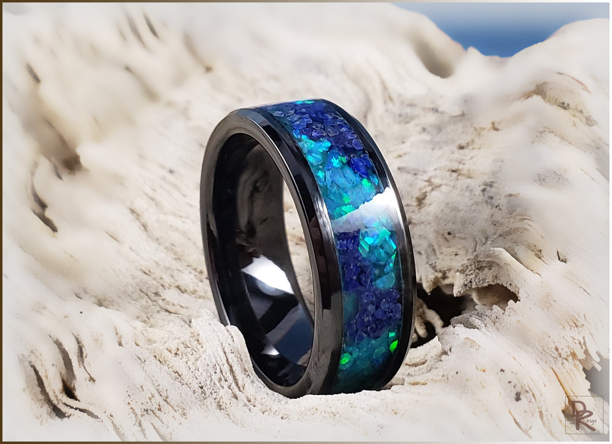 Black ceramic ring with glow inlay in blue and green hues from DR Rings displayed on textured natural surface