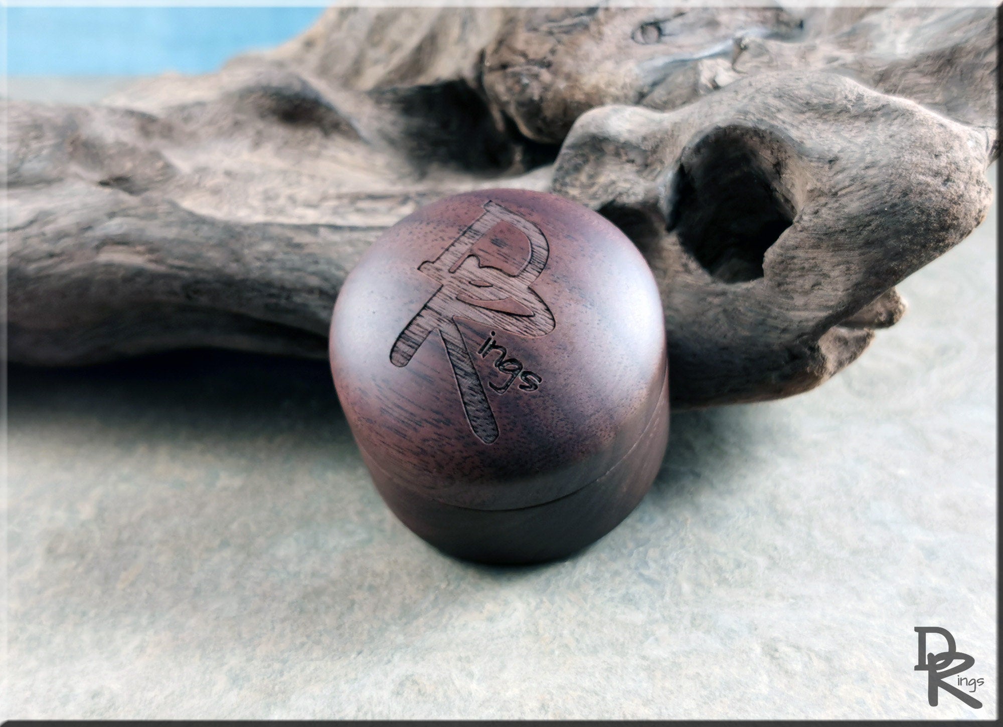 DR Rings walnut ring box with engraved logo displayed on textured surface and decorative driftwood
