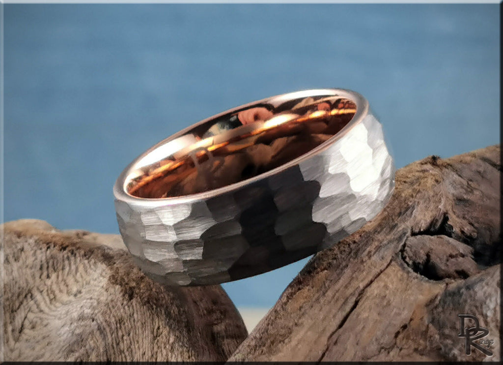Hammered tungsten ring with rose gold core resting on textured wood with blue background