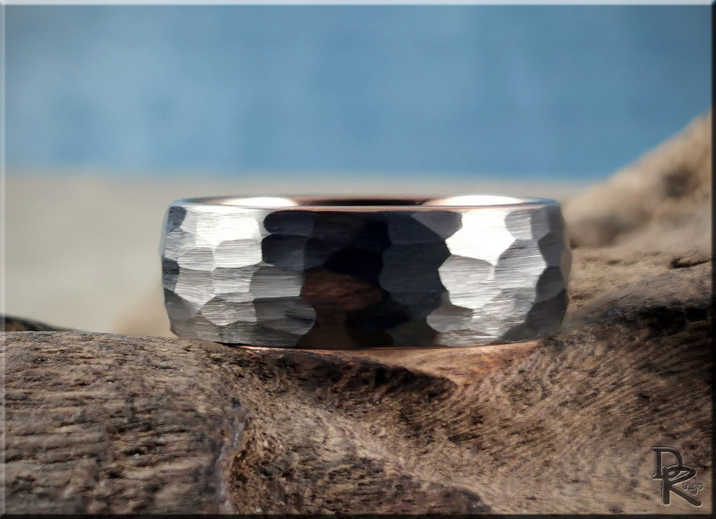 Hammered tungsten ring with rose gold core featuring a textured silver exterior on wood